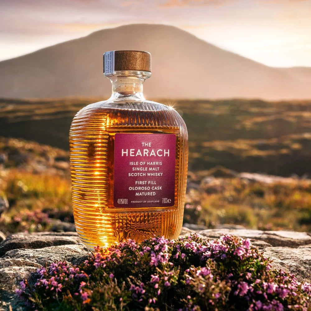 Load image into Gallery viewer, A bottle of The Hearach Oloroso Cask Single Malt Scotch Whisky stands on a rocky outcrop with purple flowers in the foreground and a mountain landscape at sunset in the background.