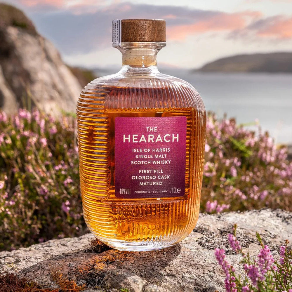 Load image into Gallery viewer, A bottle of The Hearach Oloroso Cask Single Malt Scotch Whisky stands on a rock outdoors, surrounded by pink heather flowers and a blurred coastal landscape with water and sky in the background.