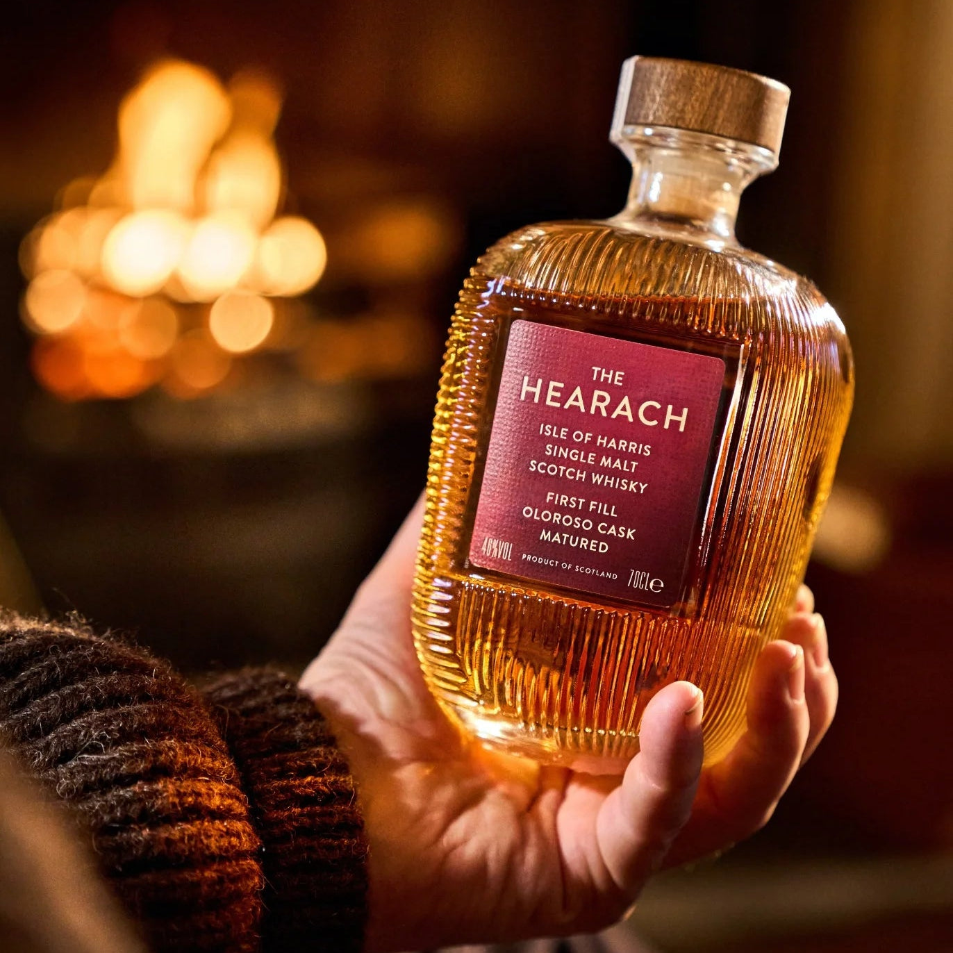Load image into Gallery viewer, A hand holds a bottle of The Hearach Oloroso Cask Single Malt Scotch Whisky against a warm, blurred background with a fire.