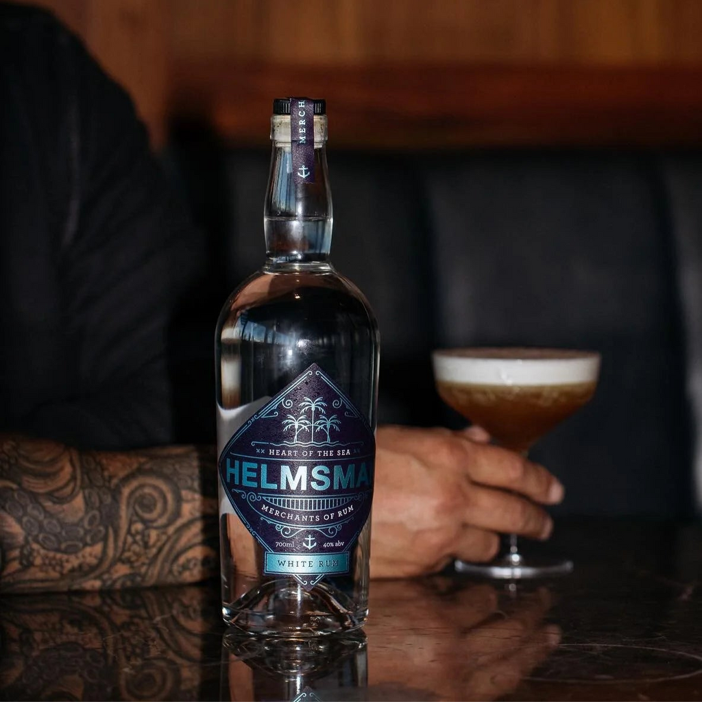 Bottle of Helmsman white rum on a bar with a cocktail in the background