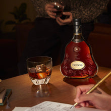 Load image into Gallery viewer, Hennessy XO bottle with a glass of whiskey on a table, person writing in a notebook.