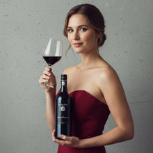 Load image into Gallery viewer, Woman in a red dress holding a bottle of Henschke Keyneton Euphonium red wine and glass of red wine against a gray background