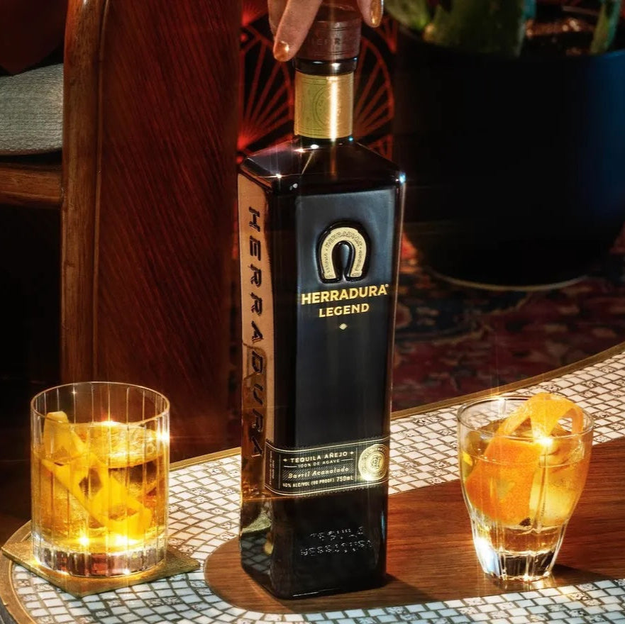 Load image into Gallery viewer, Bottle of Herradura tequila with two glasses of a yellow-orange cocktail on a wooden table.