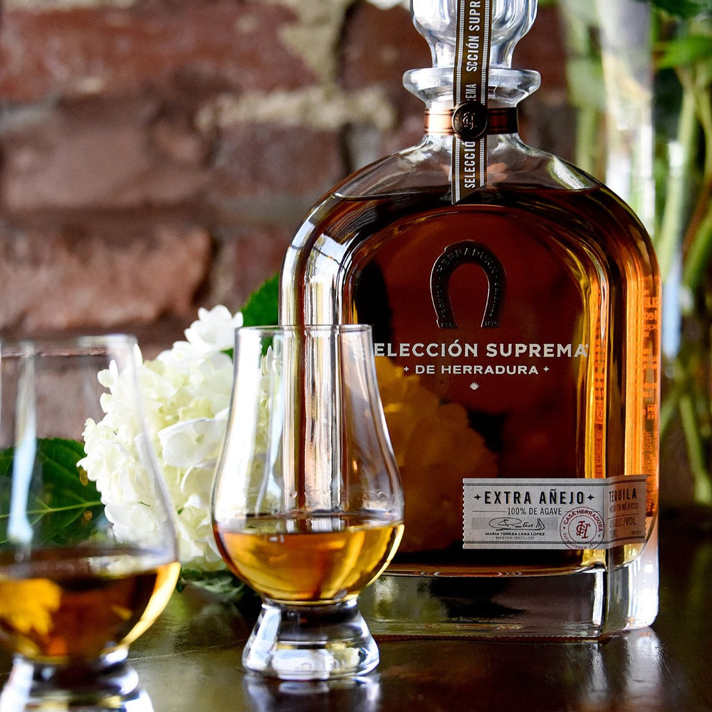 Bottle of Herradura Seleccion Supreme Extra Anejo Tequila with two glasses on a wooden surface, against a brick wall background.