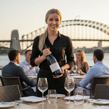 Load image into Gallery viewer, Waitress holding a bottle of Hewitson-Ned-Henry-Shiraz wine at an outdoor dining table with the Sydney Harbour Bridge in the background.