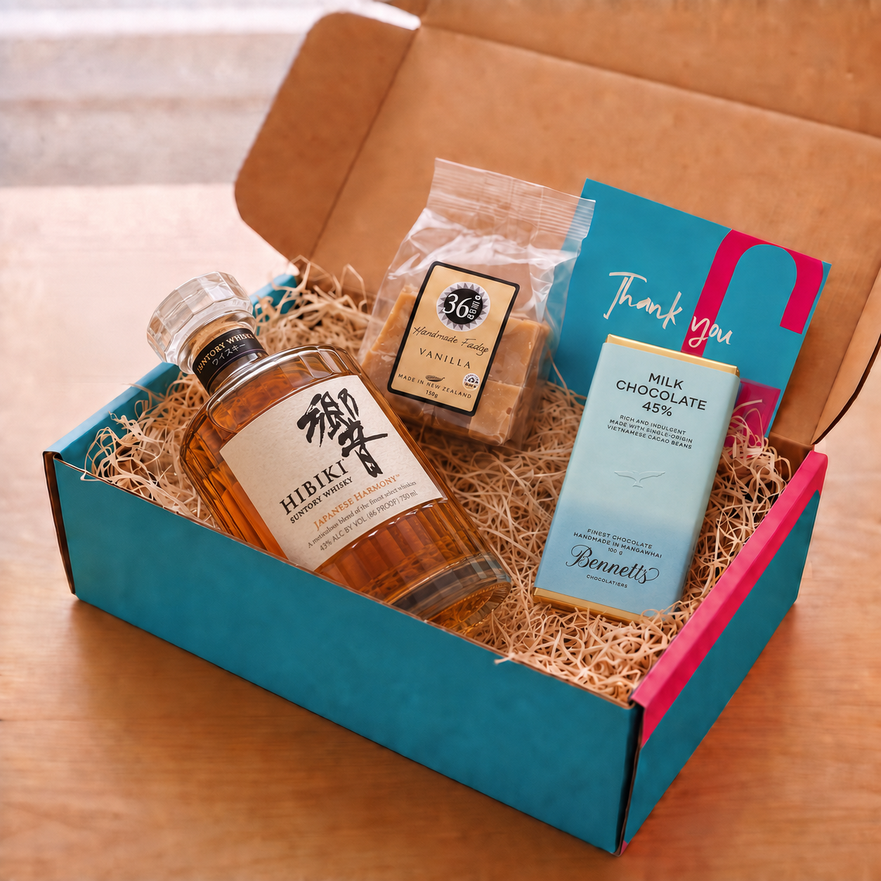 Load image into Gallery viewer, A curated gift box set on a wooden table, featuring a bottle of Hibiki Harmony Japanese Whisky, a bag of 36 Degrees Vanilla fudge, a bar of Bennett's Milk Chocolate, and a blue and pink Thank You card, all arranged in a teal and pink COVE 27 branded box with wood-wool.
