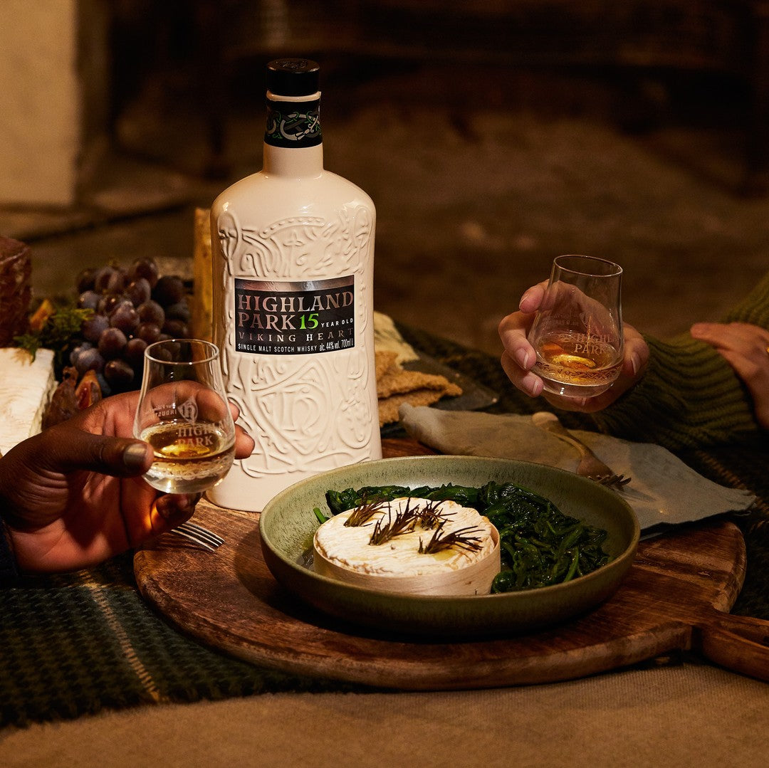 Load image into Gallery viewer, Two people holding glasses of whisky with a bottle of Highland Park whisky and a dish of food on a wooden table.
