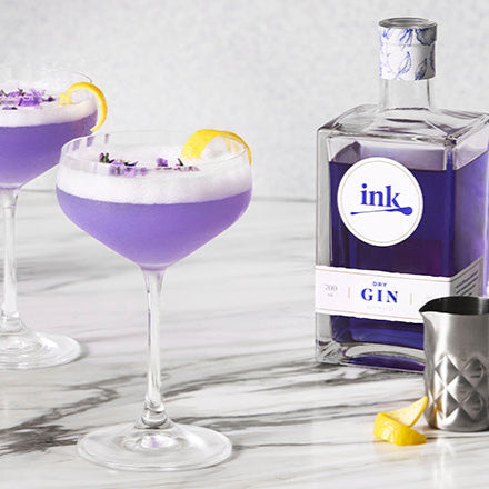 Load image into Gallery viewer, Purple cocktail in a glass with lemon twist next to a bottle of 'ink' gin on a marble surface.