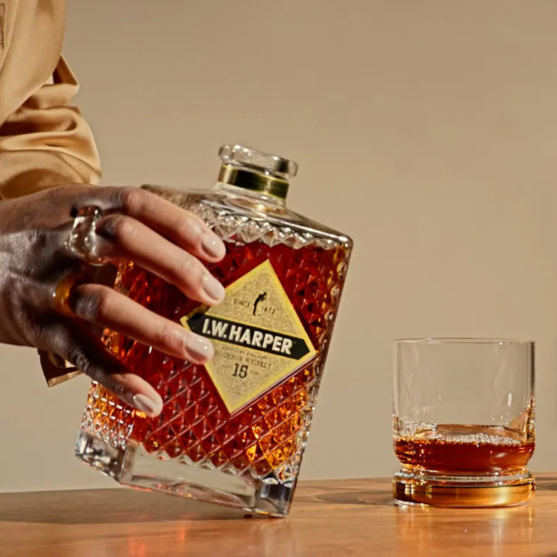 Man pouring a bottle of I.W. Harper 15 Year Old Bourbon whiskey into two glasses on a wooden surface.