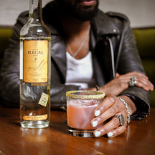 Load image into Gallery viewer, Person holding a cocktail with a bottle of Ilegal Joven Mezcal on a wooden table.