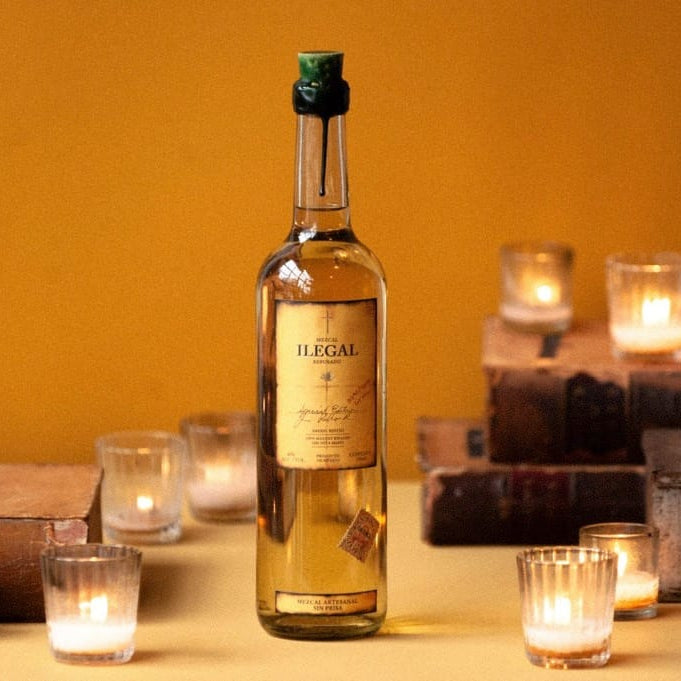 Load image into Gallery viewer, Bottle of 'Ilegal' reposado mezcal on a table with lit candles and books in the background.