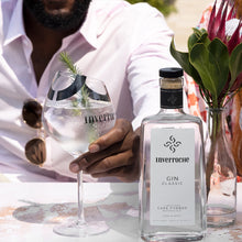 Load image into Gallery viewer, Person holding a glass of Inverroche 
gin with a bottle of Inverroche Gin Classic on a table with flowers.