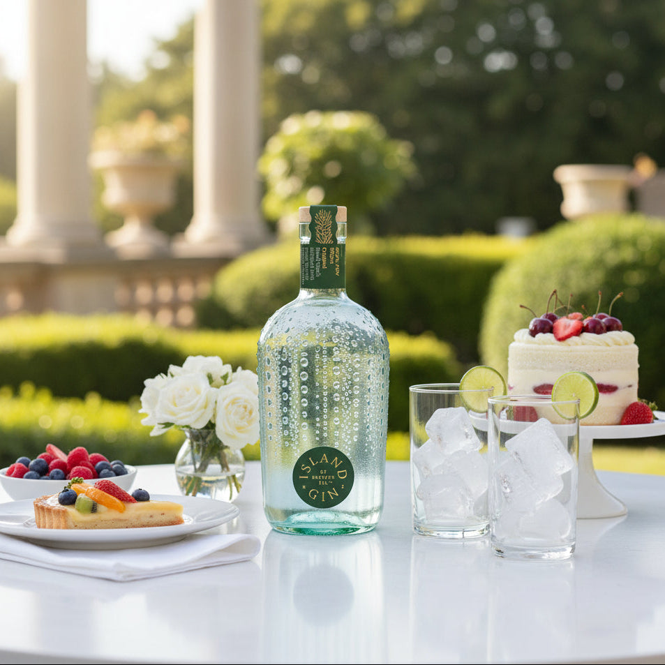 Load image into Gallery viewer, A bottle of Island Original Gin with two glasses of ice and lime slices on a white table, set against a blurred outdoor garden background with columns and a decorated cake.