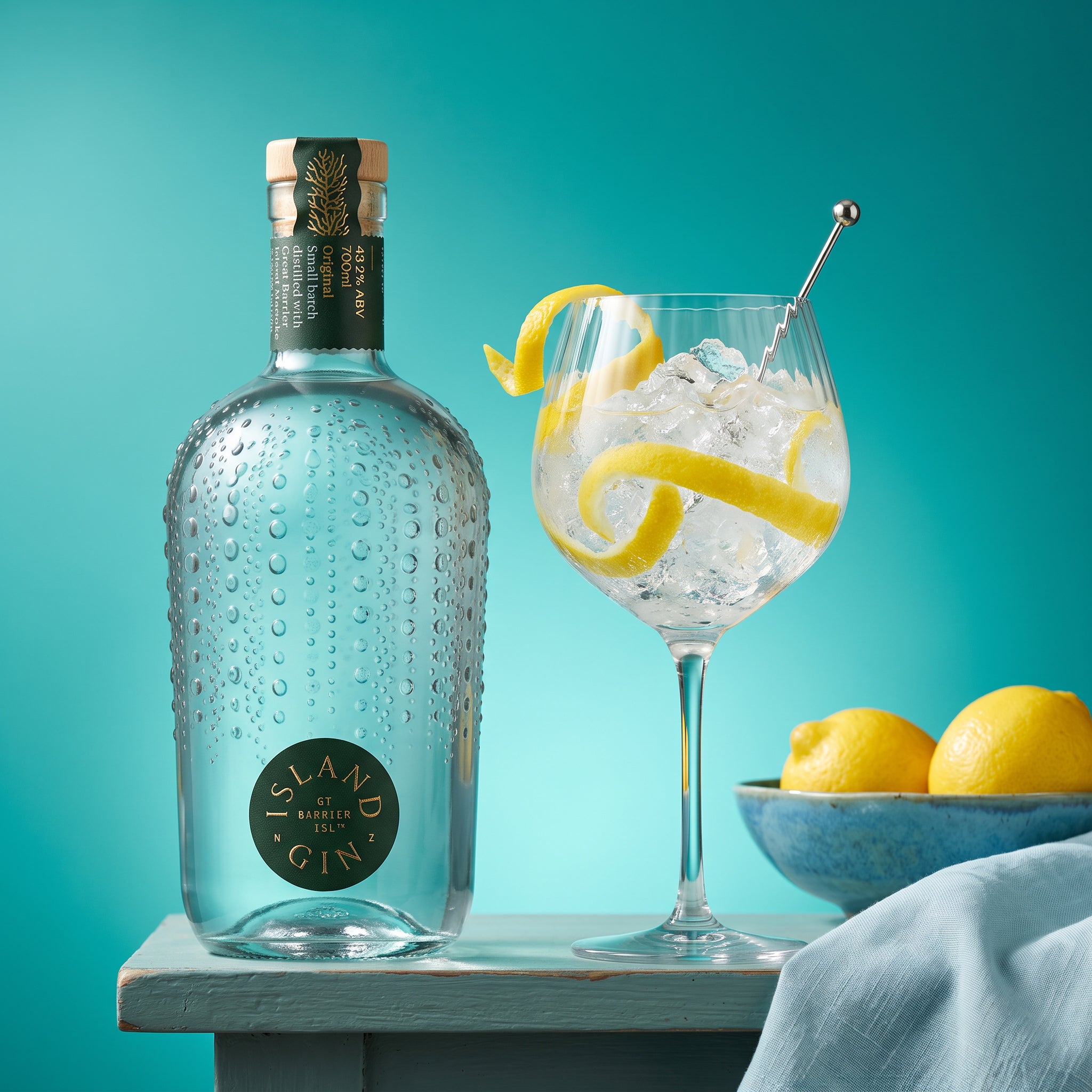 Load image into Gallery viewer, A bottle of Island Original Gin stands on a table beside a stemmed glass filled with ice and a lemon twist, with lemons in a bowl against a teal background.