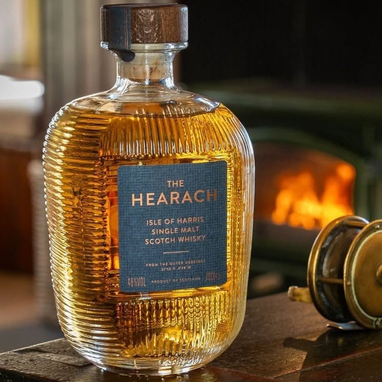 Load image into Gallery viewer, A bottle of The Hearach Isle of Harris Single Malt Scotch Whisky sits on a wooden table in a warm, cozy setting with a blurred fireplace in the background. The ribbed glass bottle is filled with golden amber whisky and features a dark blue label with gold lettering.