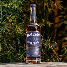 Load image into Gallery viewer, Jameson whiskey bottle on a wooden surface with green foliage in the background