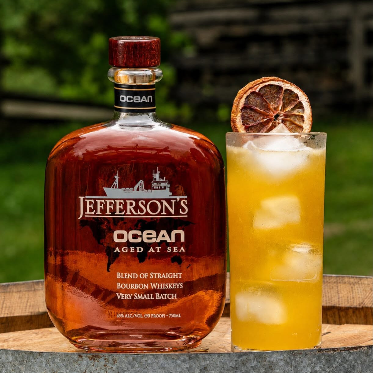 Load image into Gallery viewer, A bottle of Jefferson's Ocean Aged at Sea Bourbon Whiskey stands next to a tall glass of the coloured spirit filled with ice, garnished with a dried orange slice, set on a rustic wooden barrel outdoors with a blurred green background.