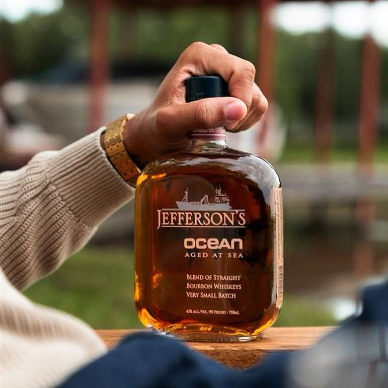 Load image into Gallery viewer, A hand holding a bottle of Jefferson's Ocean Aged at Sea bourbon whiskey on a wooden surface, with a blurred outdoor background.