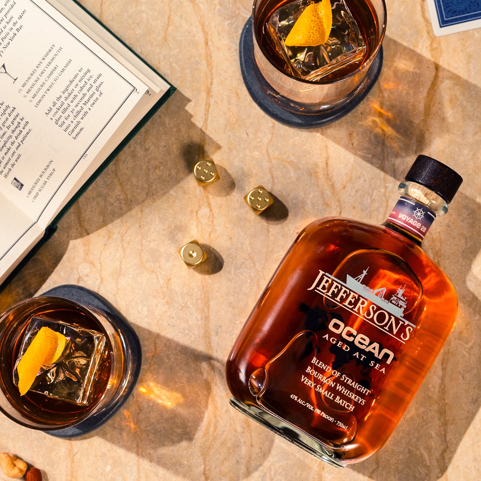Load image into Gallery viewer, A bottle of Jefferson's Ocean Aged at Sea Bourbon Whiskey is shown on a beige stone surface with a glass of the amber liquid, an ice cube, orange peel garnish, gold dice, and an open book.
