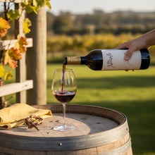 Load image into Gallery viewer, Person pouring red wine from a bottle labeled &#39;Astriglio&#39; into a glass on a wooden barrel with a vineyard background.