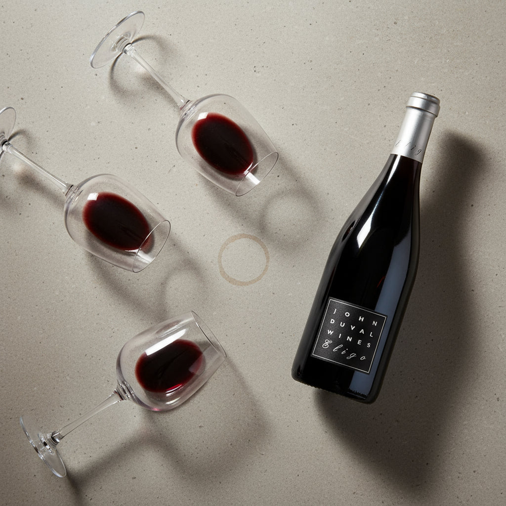 Bottle and glasses of red wine with 'John Duval' label on a light gray surface