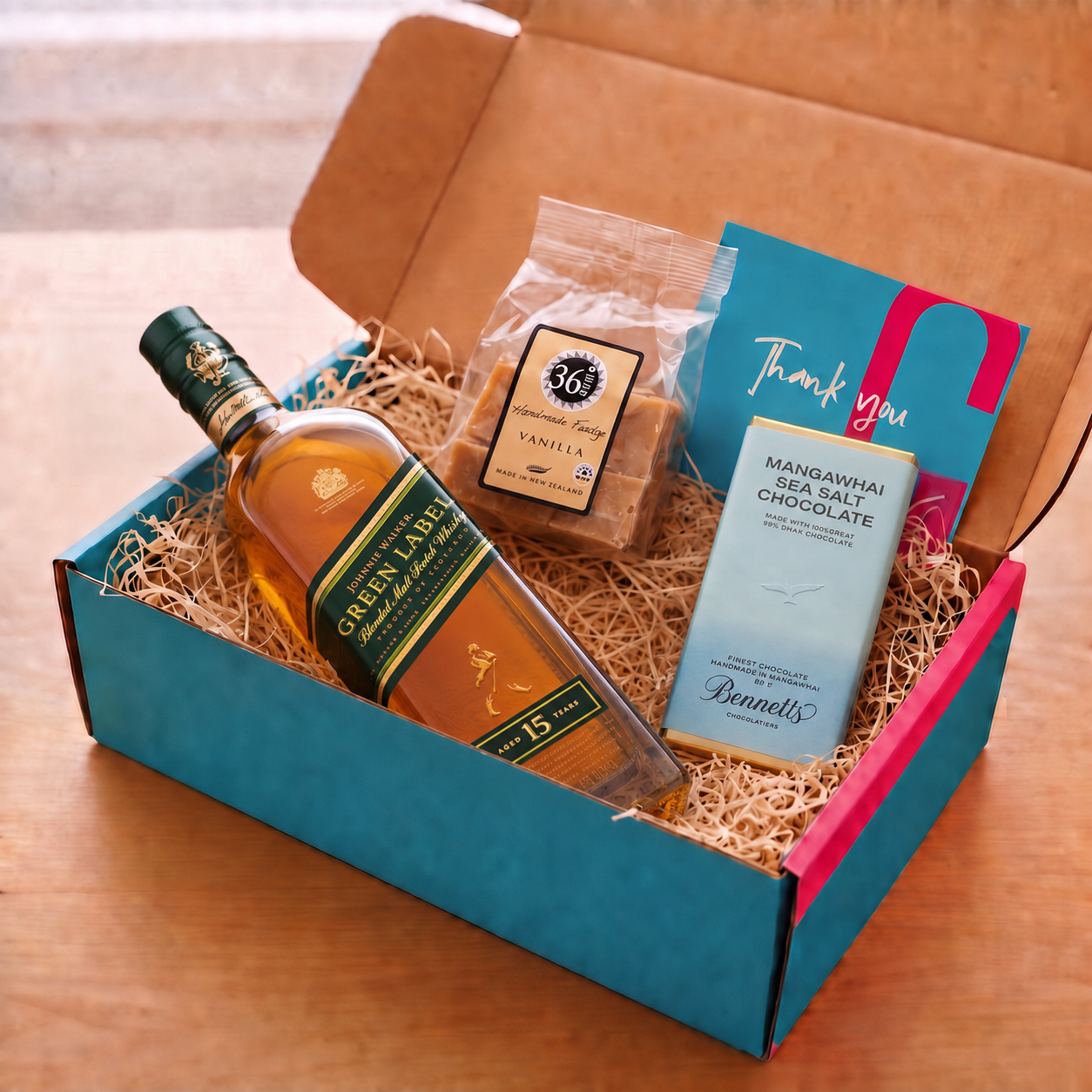 Load image into Gallery viewer, A curated gift box set on a wooden table, featuring a bottle of Johnnie Walker Green Label whisky, a bag of 36 Degrees Vanilla fudge, a blue Mangawhai Sea Salt Chocolate bar by Bennetts, and a blue 'Thank you' card inside a blue and pink box filled with wood-wool.