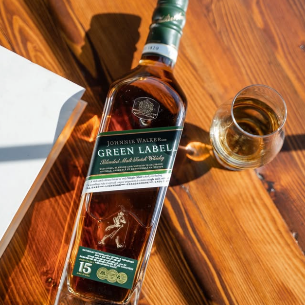 Load image into Gallery viewer, A bottle of Johnnie Walker Green Label Scotch whisky lies on a warm wooden table next to a filled glass of the amber liquid. The bottle features its signature dark green label and cap, with a white box partially visible on the left.