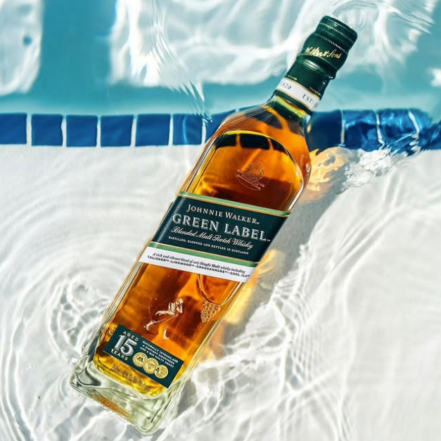 Load image into Gallery viewer, A bottle of Johnnie Walker Green Label whisky floating on rippling pool water, with blue tiled edges and bright sunlight reflecting off the glass.