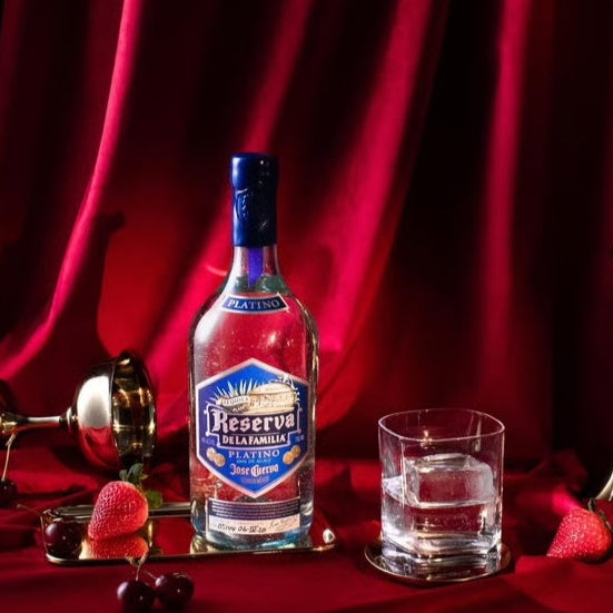 Bottle of Reserva Del La Familia tequila on a tray with a glass of water and decorative elements against a red curtain background.