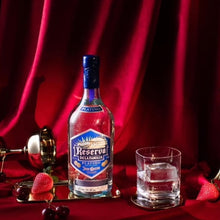 Load image into Gallery viewer, Bottle of Reserva Del La Familia tequila on a tray with a glass of water and decorative elements against a red curtain background.