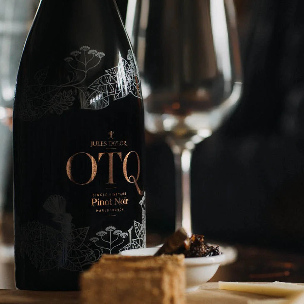 Bottle of Jules Taylor OTQ Pinot Noir with a wine glass and snacks on a table.