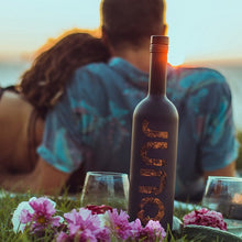 Load image into Gallery viewer, Couple sitting by a lake with a bottle of Juno Fine Gin and glasses on a grassy area.