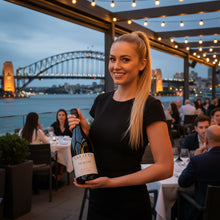 Load image into Gallery viewer, Woman holding a bottle of Kaesler The Bogan Shiraz wine with a Sydney Australia cityscape and harbour bridge in the background
