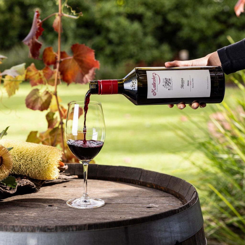 Person pouring Kalleske-Moppa-Shiraz red wine from a bottle into a glass on a wooden barrel with autumn leaves in the background.