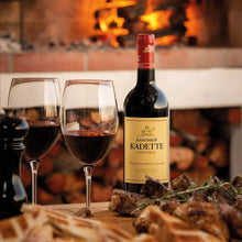 Load image into Gallery viewer, Kanonkop Kadette Pinotage wine bottle with two glasses and food on a table in front of a fireplace.