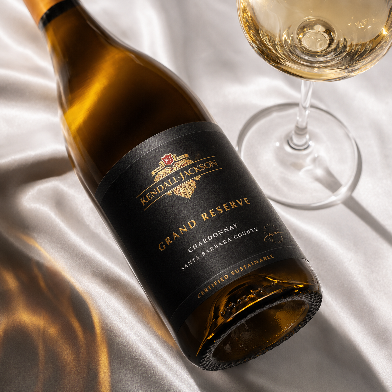 Load image into Gallery viewer, A bottle of Kendall Jackson Grand Reserve Chardonnay wine lies on draped white satin fabric next to a filled wine glass. The bottle has a black label with gold lettering, and the scene is lit with warm, directional light.
