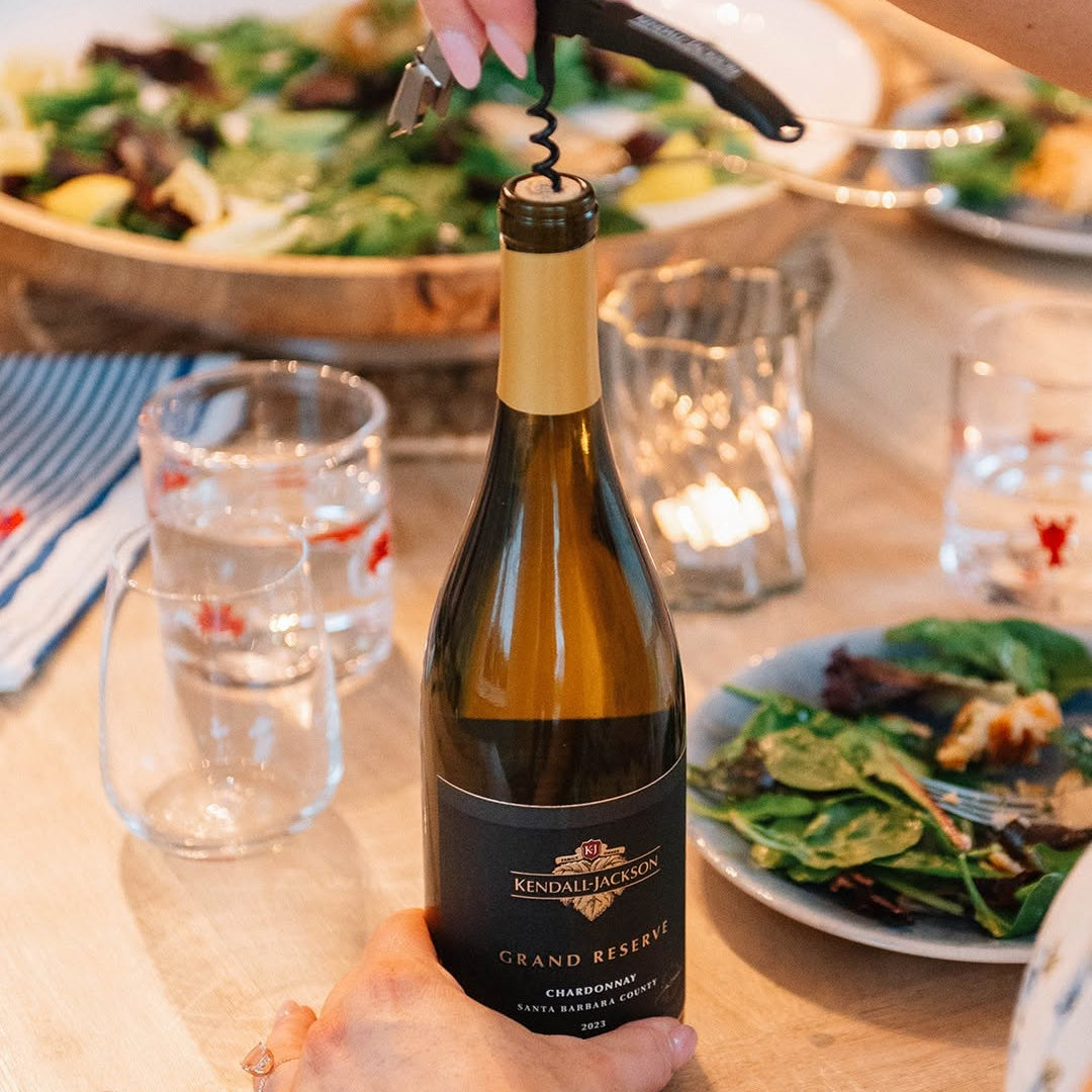 Load image into Gallery viewer, A bottle of Kendall-Jackson Grand Reserve Chardonnay being opened with a corkscrew on a dining table, surrounded by plates of salad and glasses.