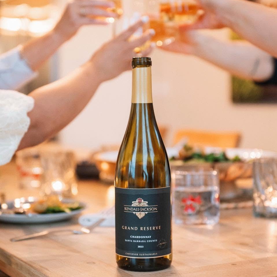 Load image into Gallery viewer, A bottle of Kendall Jackson Grand Reserve Chardonnay sits on a wooden table in a warmly lit dining setting, with multiple people's hands holding wine glasses raised in a toast in the background.