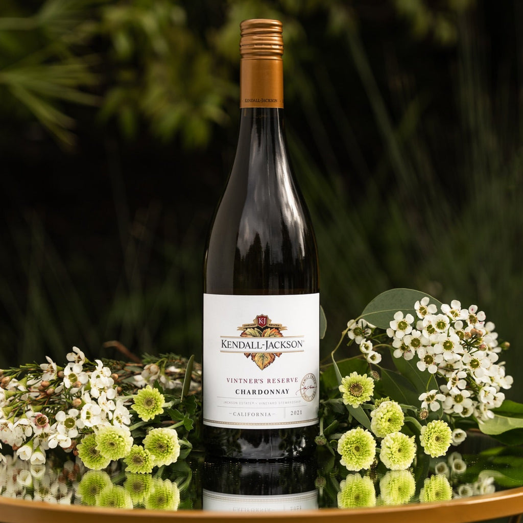 Bottle of Kendall-Jackson Chardonnay surrounded by flowers on a reflective surface with a blurred green background