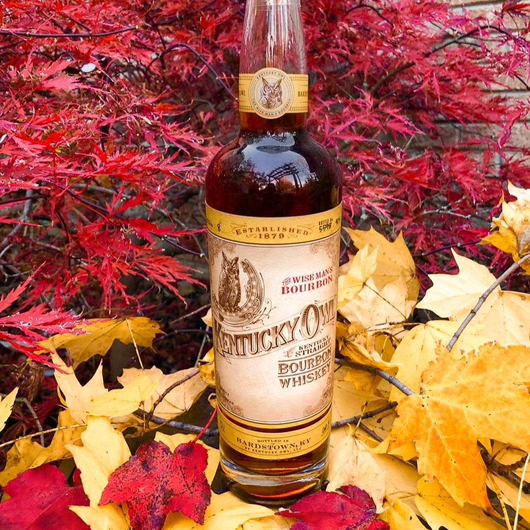 Load image into Gallery viewer, Bottle of Kentucky Owl Bourbon whiskey surrounded by autumn leaves in front of a brick wall.