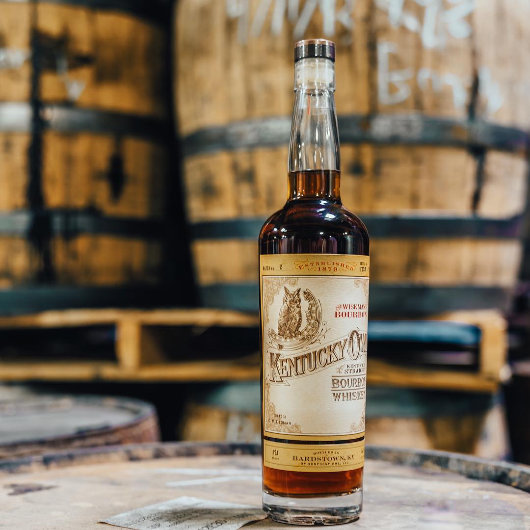 Load image into Gallery viewer, Bottle of Kentucky Owl bourbon whiskey in front of wooden barrels.