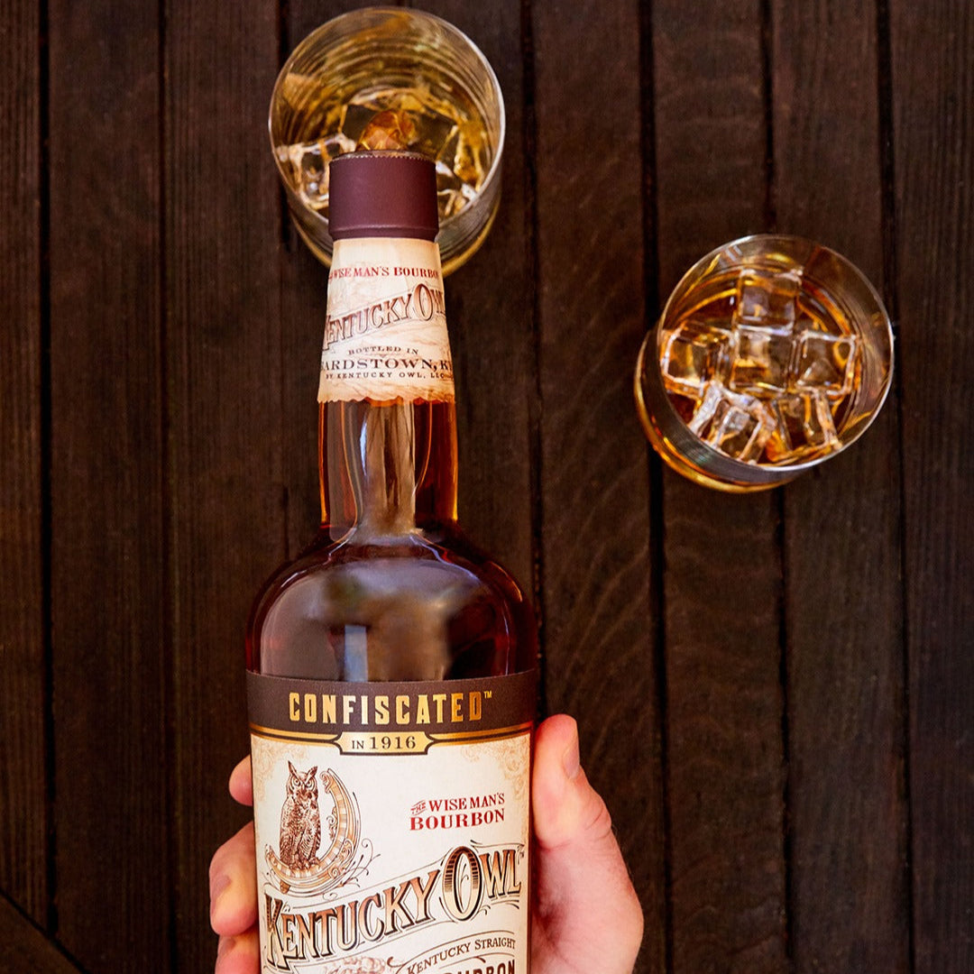 Load image into Gallery viewer, Bottle of Kentucky Owl bourbon whiskey held by a hand with two glasses of whiskey in the background.