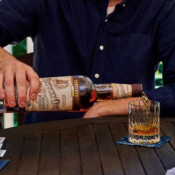 Load image into Gallery viewer, Person pouring whiskey from a bottle into a glass on a wooden table.