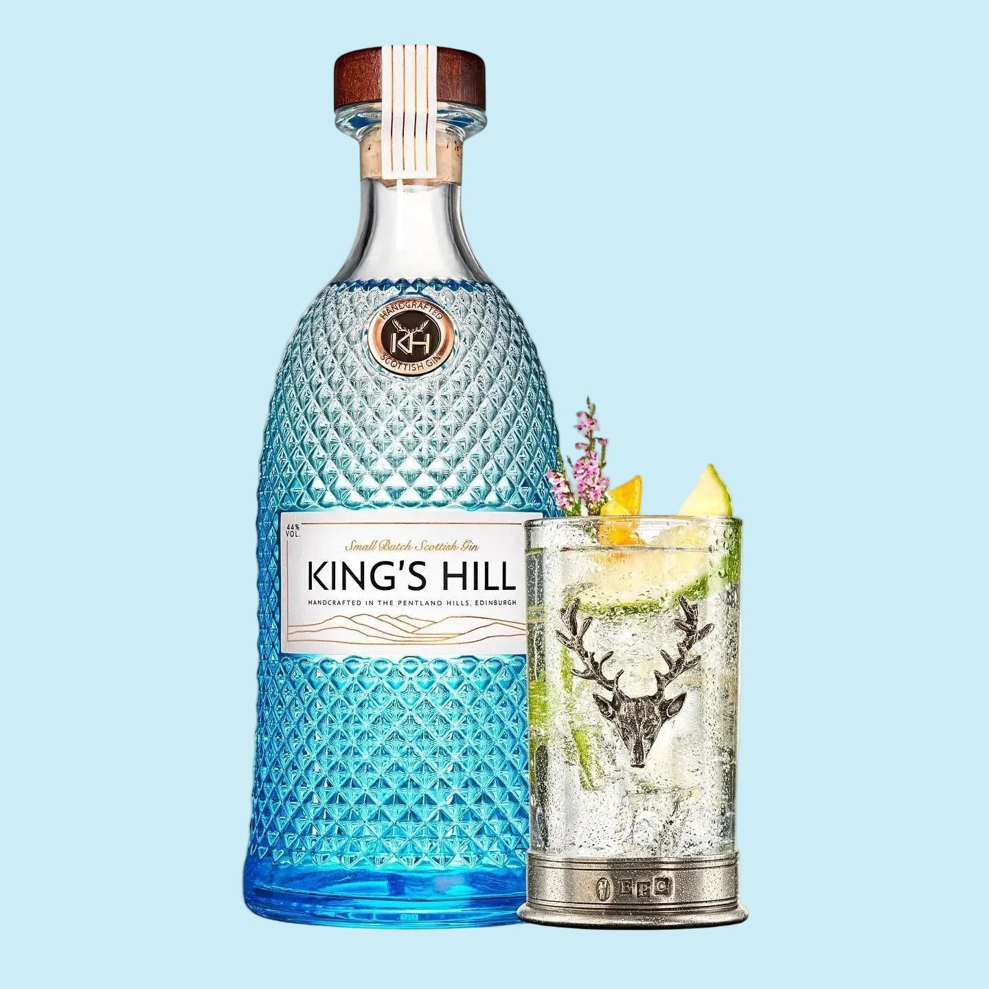 Load image into Gallery viewer, King's Hill Gin bottle in a textured blue glass with a wooden stopper, shown next to a clear cocktail glass filled with a clear gin and garnished with lime and a deer decanter, on a white background.