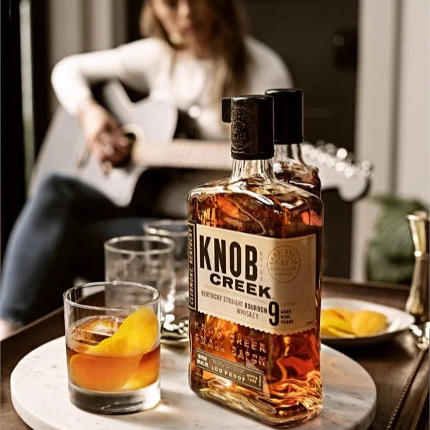 Bottle of Knob Creek whiskey with a glass on a table as a person plays guitar in the background.