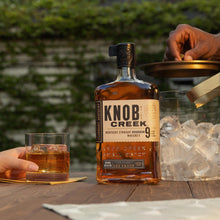 Load image into Gallery viewer, Knob Creek 9 year old whiskey bottle on a wooden table with a glass of whiskey and ice cubes.