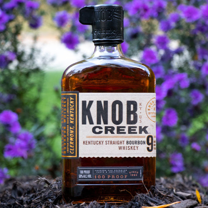 Load image into Gallery viewer, Bottle of Knob Creek bourbon whiskey with purple flowers in the background.