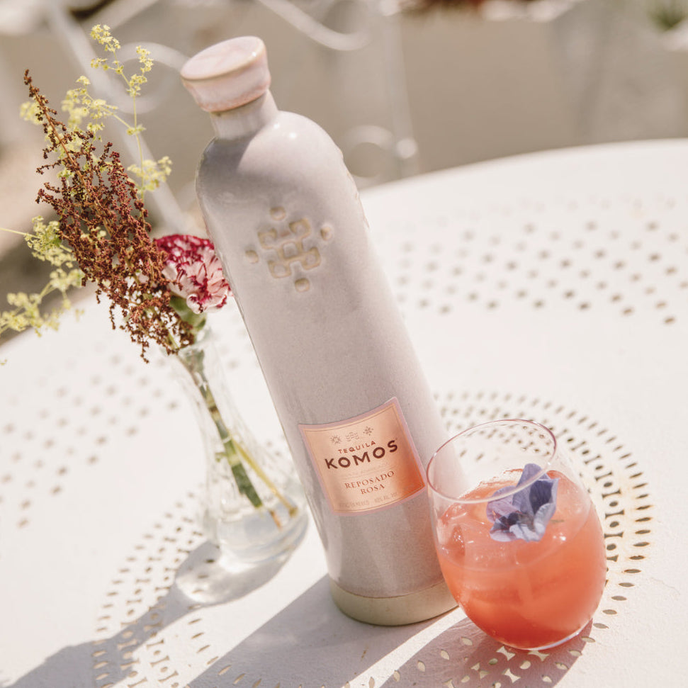 Load image into Gallery viewer, A Bottle of Komos Reposado Rosa Tequila next to a pink cocktail on a white table outdoors.