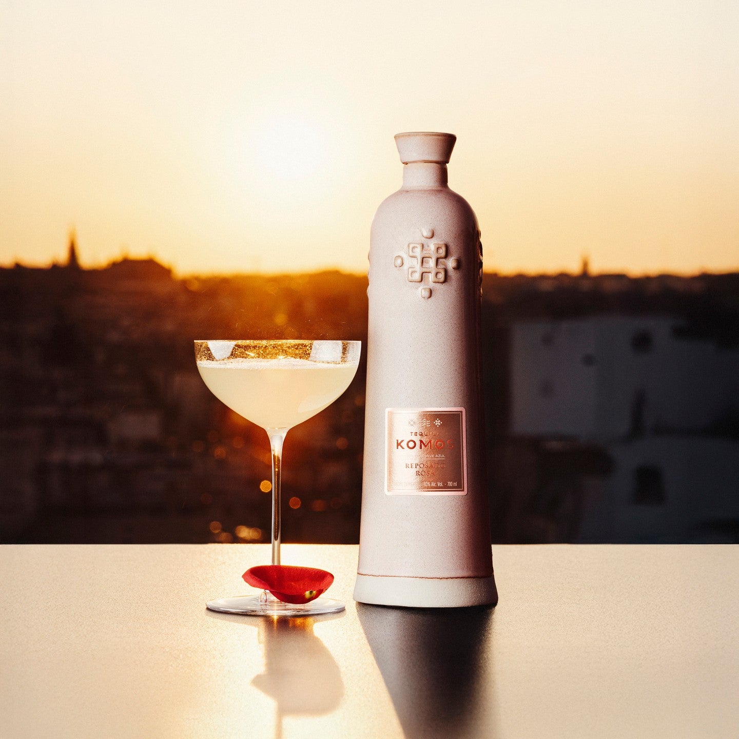 Load image into Gallery viewer, Bottle of Komos Reposado Rosa Tequila with a cocktail glass against a sunset backdrop