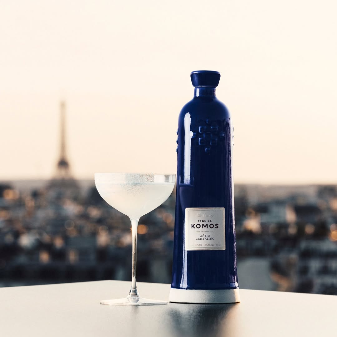Load image into Gallery viewer, Bottle of Komos Añejo Cristalino Tequila next to a cocktail glass against a blurred Paris cityscape background